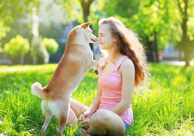 Woman with pink top on sits on the ground in a park while her dog sniffs her face.